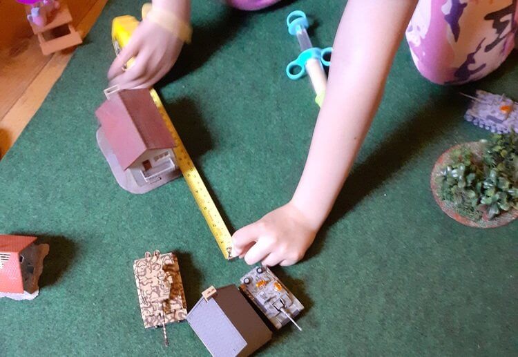 Photo of a Panzer Kids game. A player is using a tape measure to measure a model StuG's movement. Also visible are a model Tiger tank, buildings and a wood.